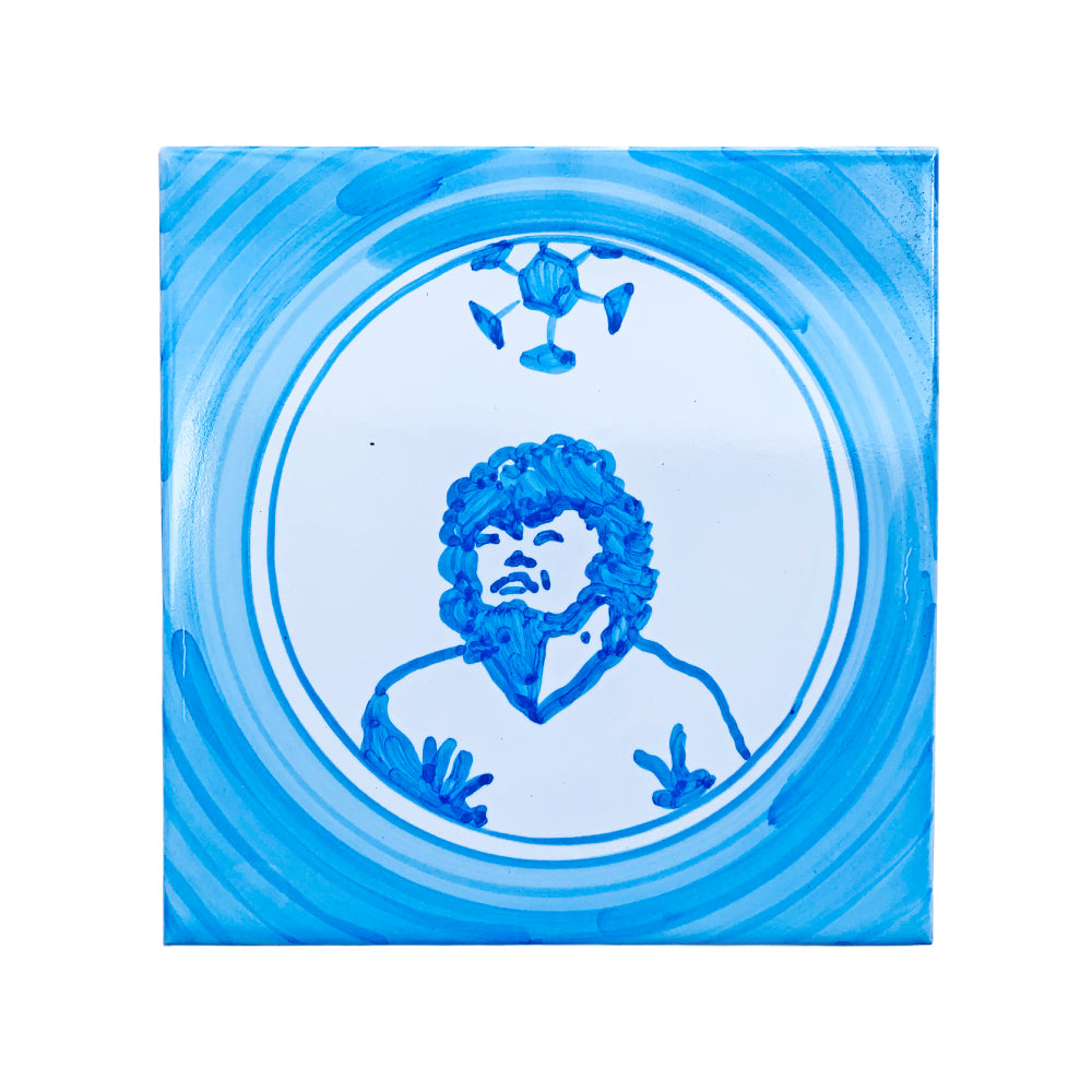 Maiolica decorativa Pil Pil con illustrazione in blu di Diego Maradona, El Pibe de Oro, stilizzato in stile tradizionale, con pallone sopra la testa e cornice circolare su sfondo bianco e azzurro.