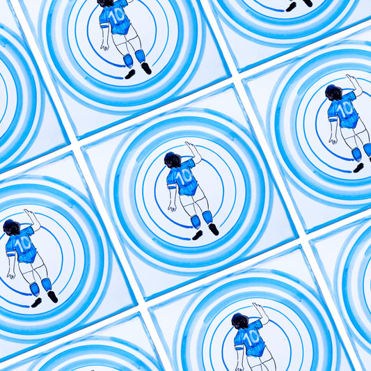 Maiolica Pil Pil raffigurante Diego Armando Maradona di spalle con la maglia numero 10, immerso in cerchi concentrici azzurri su sfondo bianco, in stile pop iconico napoletano.