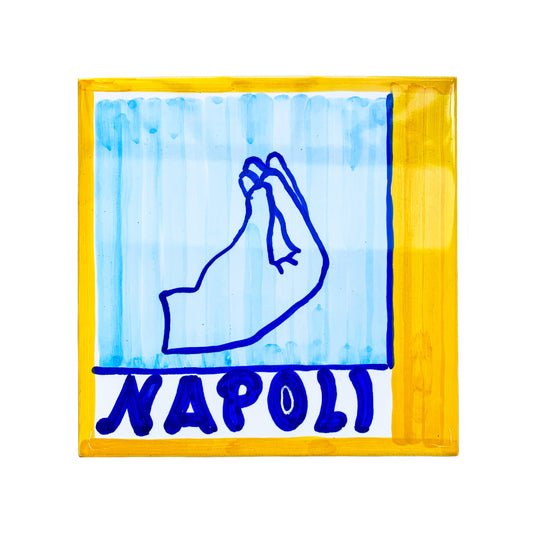 Maiolica artigianale del brand Pil Pil, dipinta a mano e realizzata in terracotta di pasta rossa smaltata. La piastrella, di dimensioni 20x20 cm, presenta un bordo giallo brillante e uno sfondo azzurro decorato con un disegno stilizzato di una mano che compie il gesto napoletano del ma che vuoi, simbolo di espressivita e cultura partenopea. Nella parte inferiore spicca la scritta Napoli in blu intenso.